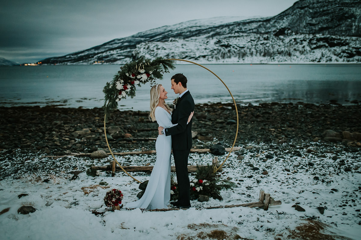 Bride and groom reading their vows to each other under a round floral arch in front of a winter and snowy landscape by the fjord in Alta Norway