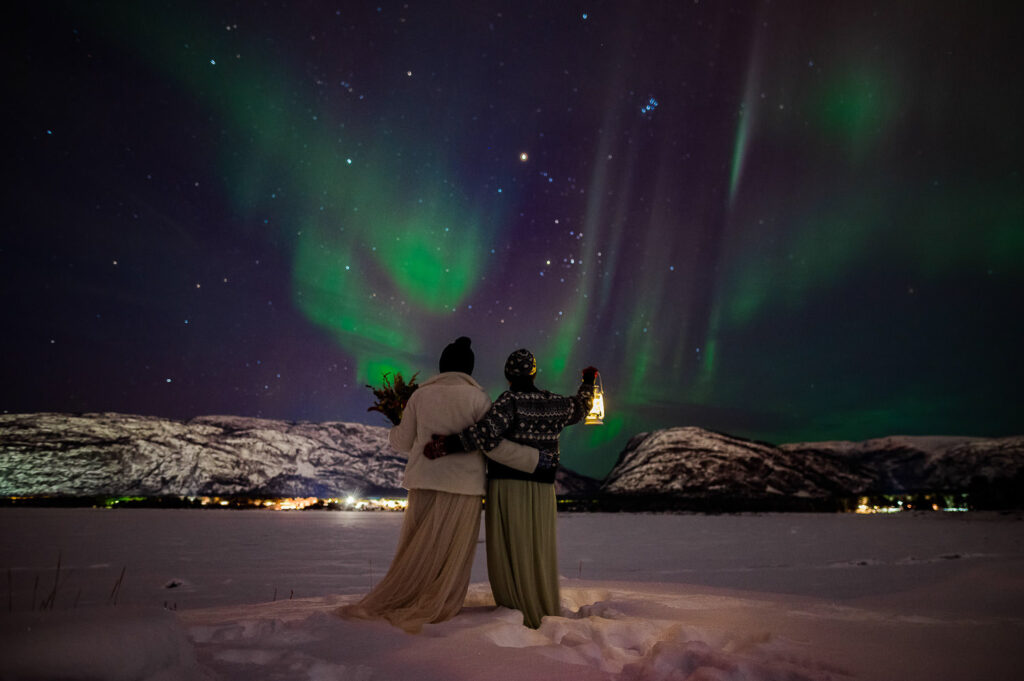 Two brides in wedding dresses hugging each other while watching beautiful northern lights on the day of their LGBT elopement under Aurora Borealis in Alta Norway