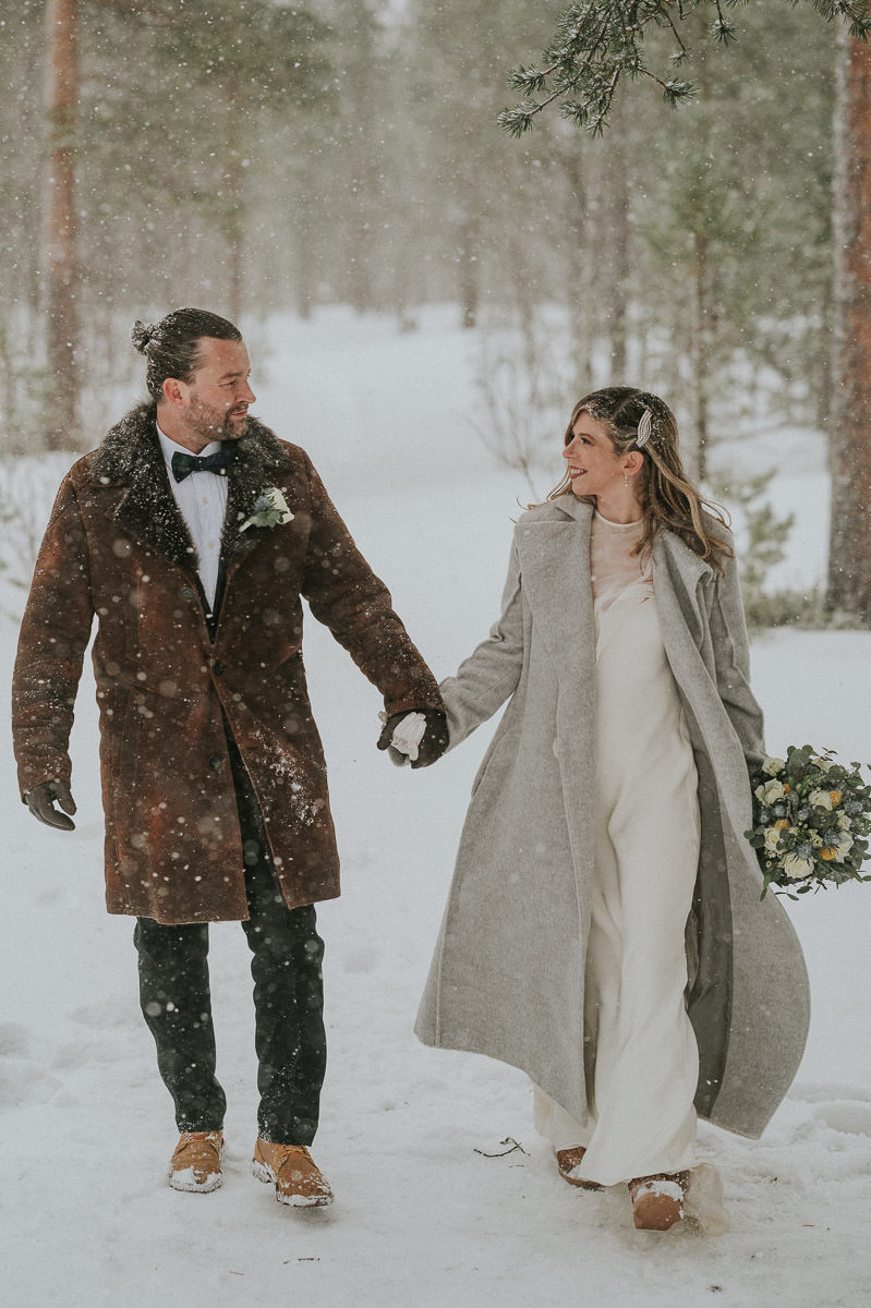 Cute couple - bride and groom in their wedding attire and with winter coats on walking hand in hand in a forest under a snowfall