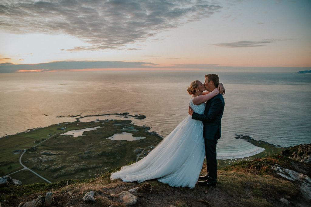 Brudepar kysser hverandre på en fjelltopp i Lofoten med en nydelig solnedgang i bakgrunn