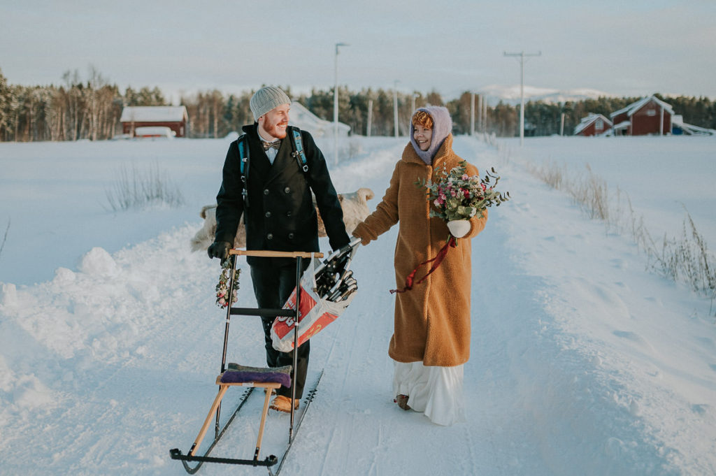 Bride and groom walking to their elopement location on the day of their winter elopement in Northern Norway, the groom is kicking a kicksled in front of him and he is carrying snowshoes