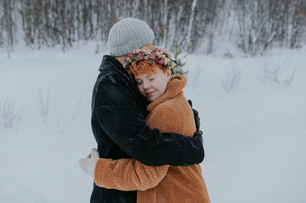 Groom is hugging his bride to warm her up on a cold winter day of their wedding in Norway, The bride is wearing a beautiful floral crown of pink and blush flowers