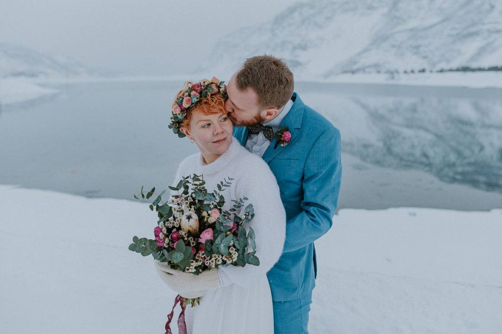 Groom kissing his bride on a cheek on the day of their outdoor winter elopement in Norway - with a beautiful winter fjord and mountain landscape in the background