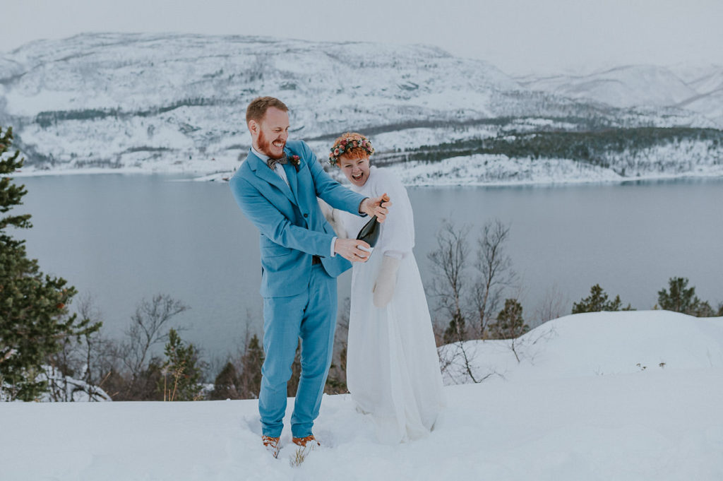 Eloping couple popping up champagne in front of beautiful mountain and fjords landscapes in Northern Norway 