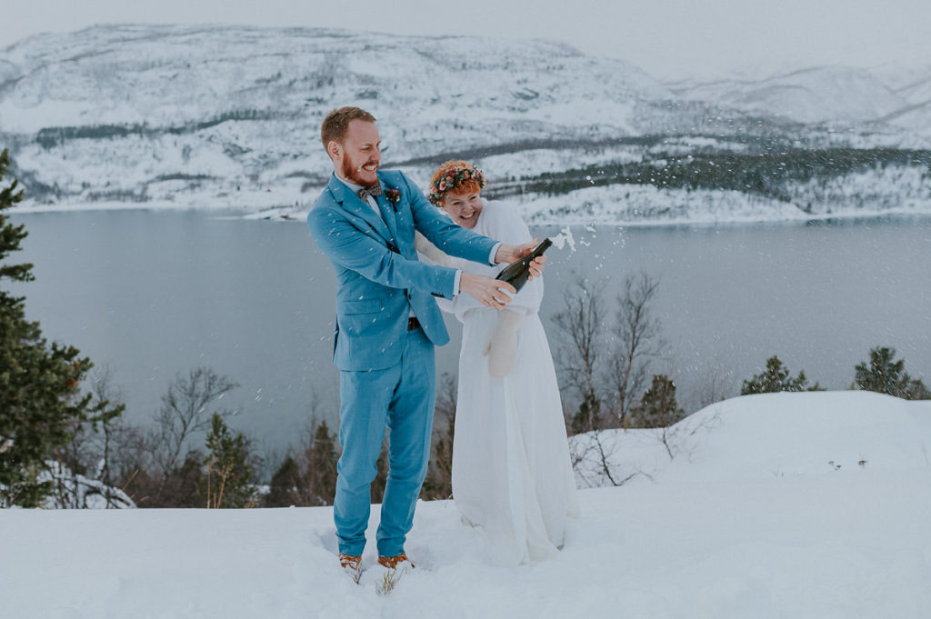 Eloping couple popping up prosecco bottle in front of beautiful mountain and fjords landscapes in Northern Norway in winter time