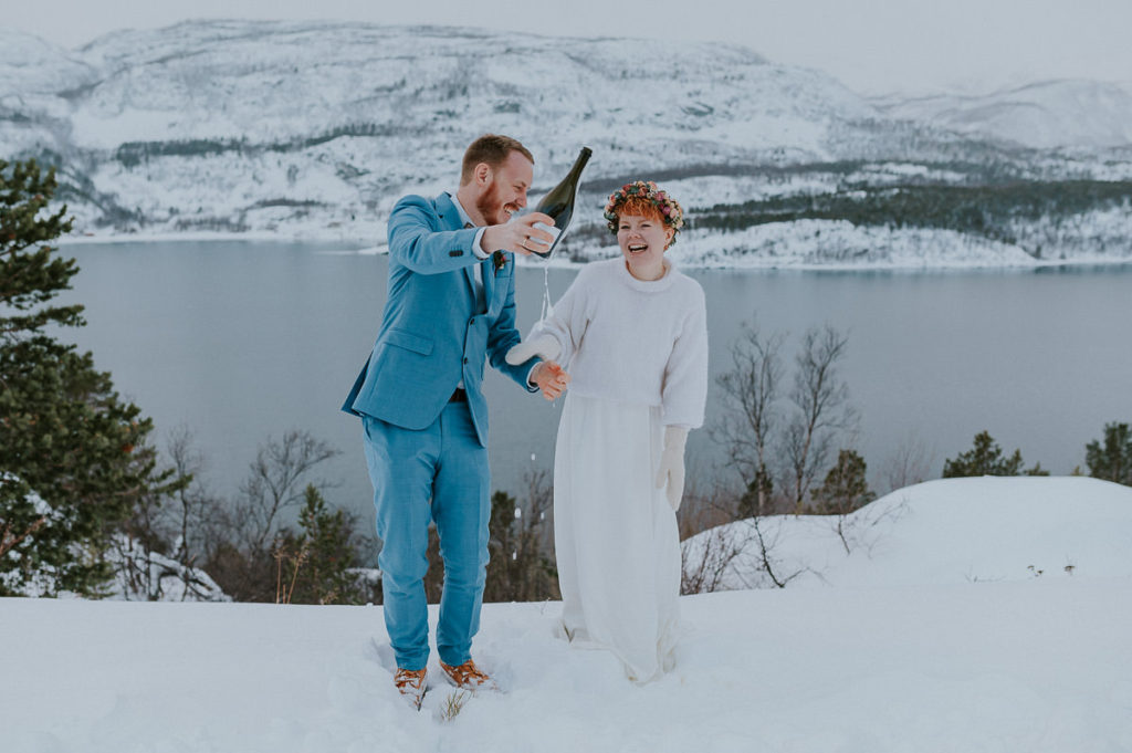 Eloping couple popping up sparkling wine in front of beautiful mountains and fjords in Northern Norway 