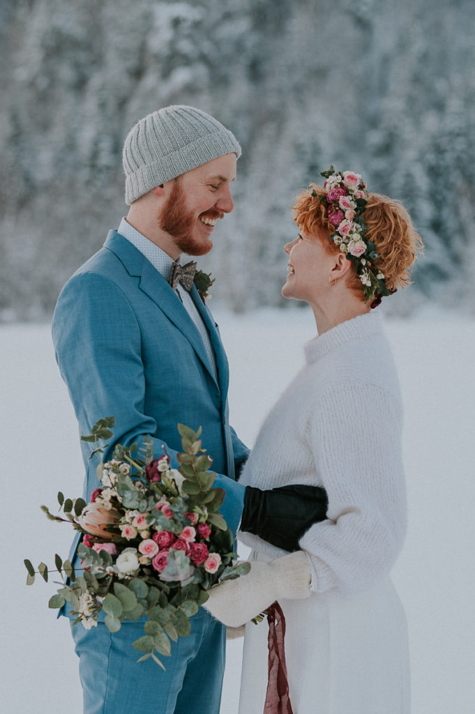 Bride and groom in front of winter forest in Alta Norway both smiling and looking happy. Bride wears flower crown in pink color and holding a gorgeous bouquet 