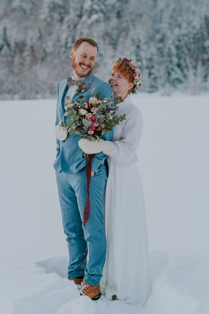 Bride and groom in front of winter forest in Alta Norway both smiling and looking happy. Bride wears flower crown in pink color and holding a gorgeous bouquet - captured by Alta Norway elopement photographer TS Foto Design
