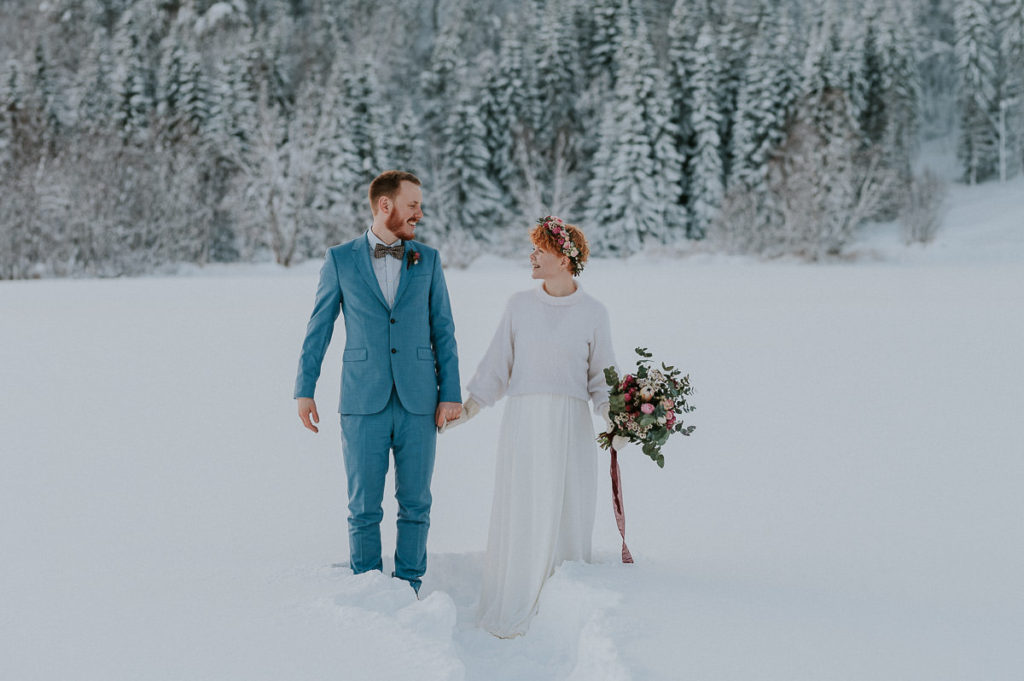 Bride and groom in front of winter forest in Alta Norway both smiling and looking happy. Bride wears flower crown in pink color and holding a gorgeous bouquet - captured by Alta Norway elopement wedding  photographer TS Foto Design