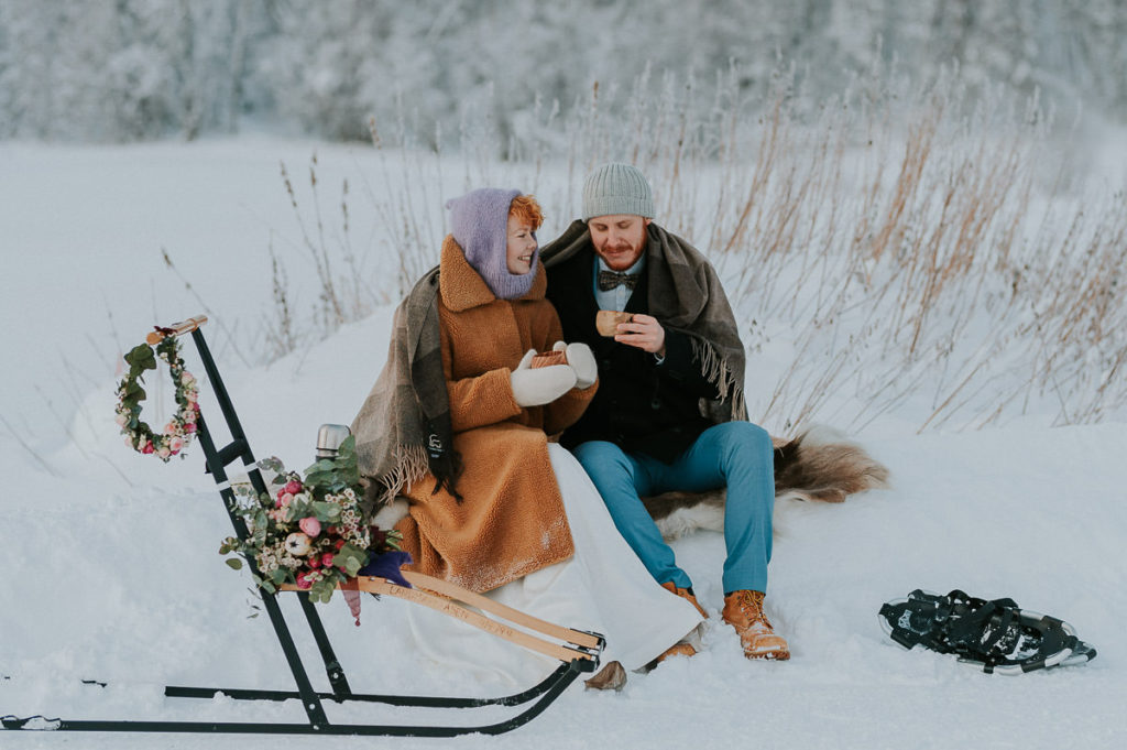 Bride and groom drinking coffee outdoors in the winter landscape of Alta in Norway. They are wearing warm wool coats and beanies and are properly prepared for their outdoor wedding in Northern Norway