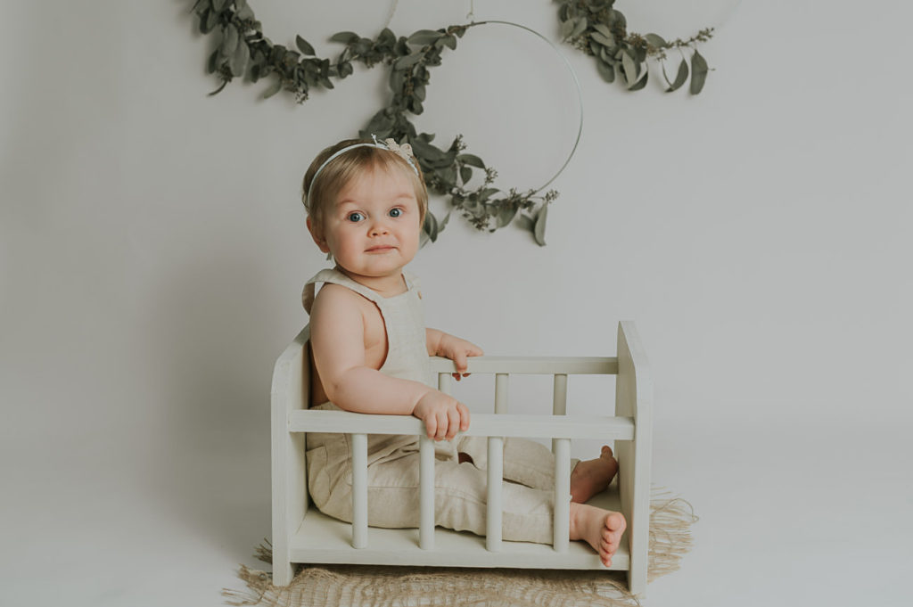 Milepælfotografering babyfoto i Alta i fotostudio TS Foto Design. Skjønn liten jente sitter i en dukkeseng med eucalyptus pynt i bakgrunn