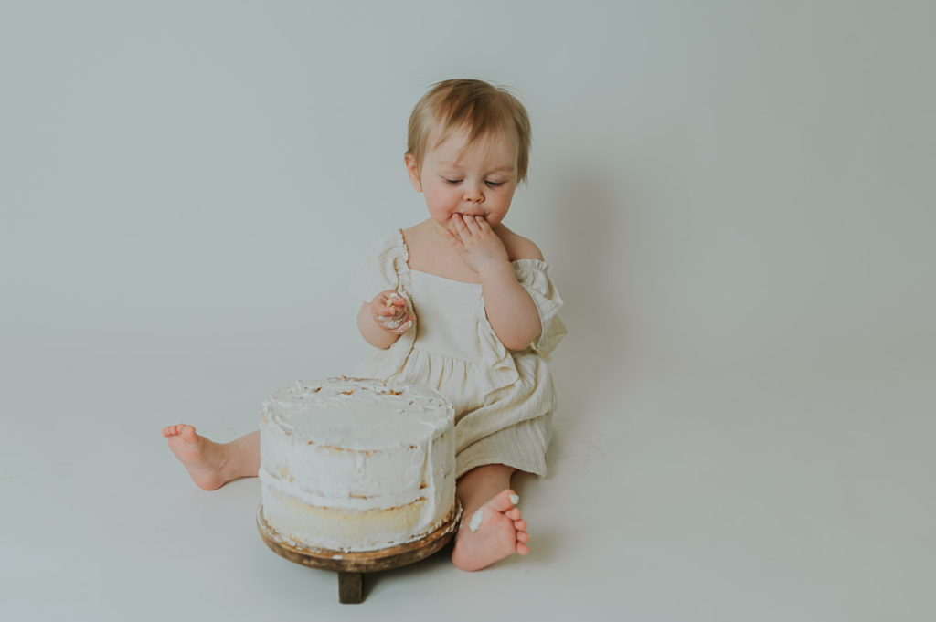 Ettårsfotografering cake smash for en baby jente - babyen smaker på kaken sin med hvit foto bakgrunn