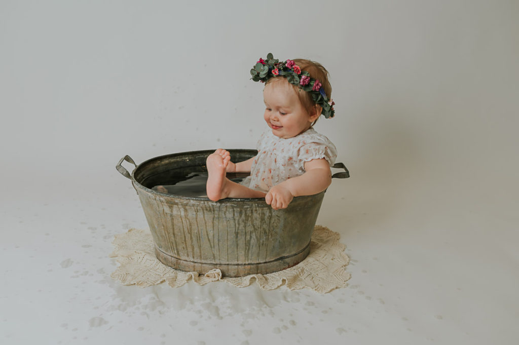 Ettårsfotografering i fotostudio i Alta TS Foto Design. Babyen sitter i en sinkbalje med en fin blomsterkrans på hodet. Hun plasker og smiler og er så fornøyd