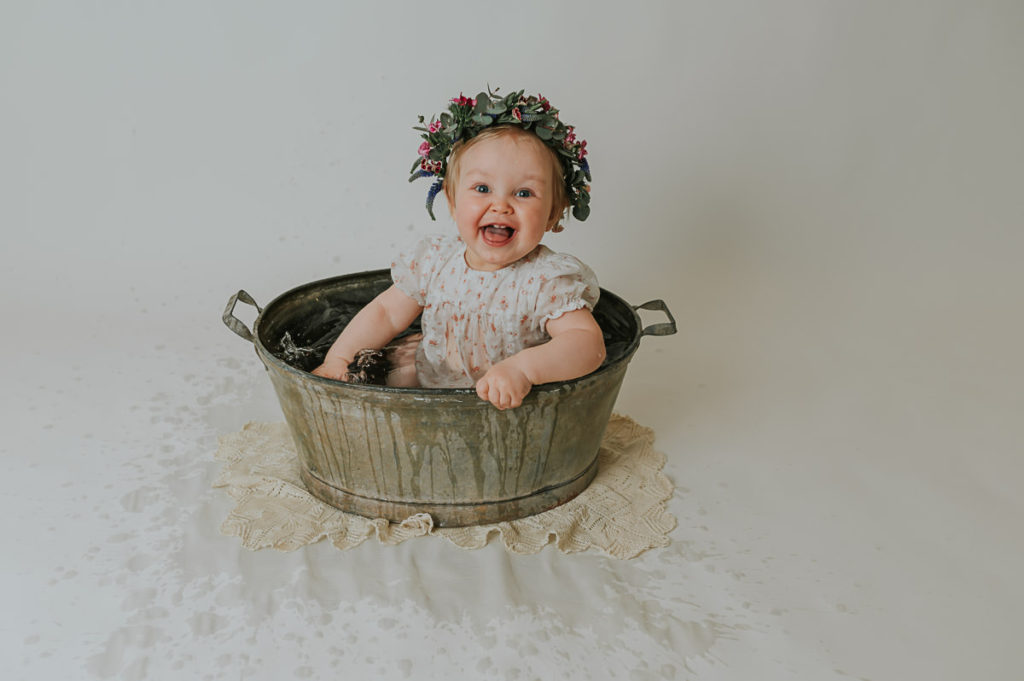 Ettårsfotografering i fotostudio i Alta TS Foto Design. Babyen sitter i en sinkbalje med en fin blomsterkrans på hodet. Hun plasker og smiler og er så fornøyd