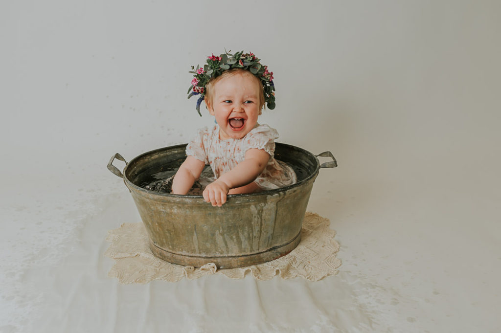 Barnefotografering i fotostudio i Alta TS Foto Design. Ett år gammel baby jente sitter i en sinkbalje med en fin blomsterkrans på hodet. Hun plasker og smiler og er så fornøyd