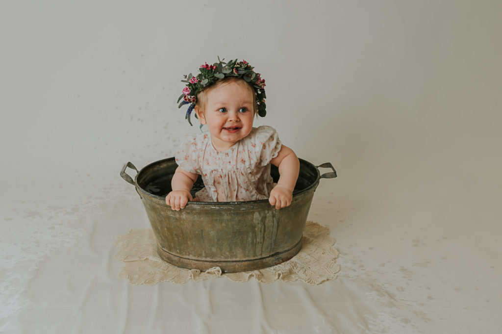 Ettårsfoto i fotostudio i Alta TS Foto Design. Babyen sitter i en sinkbalje med en fin blomsterkrans på hodet. Hun plasker og smiler og er så fornøyd