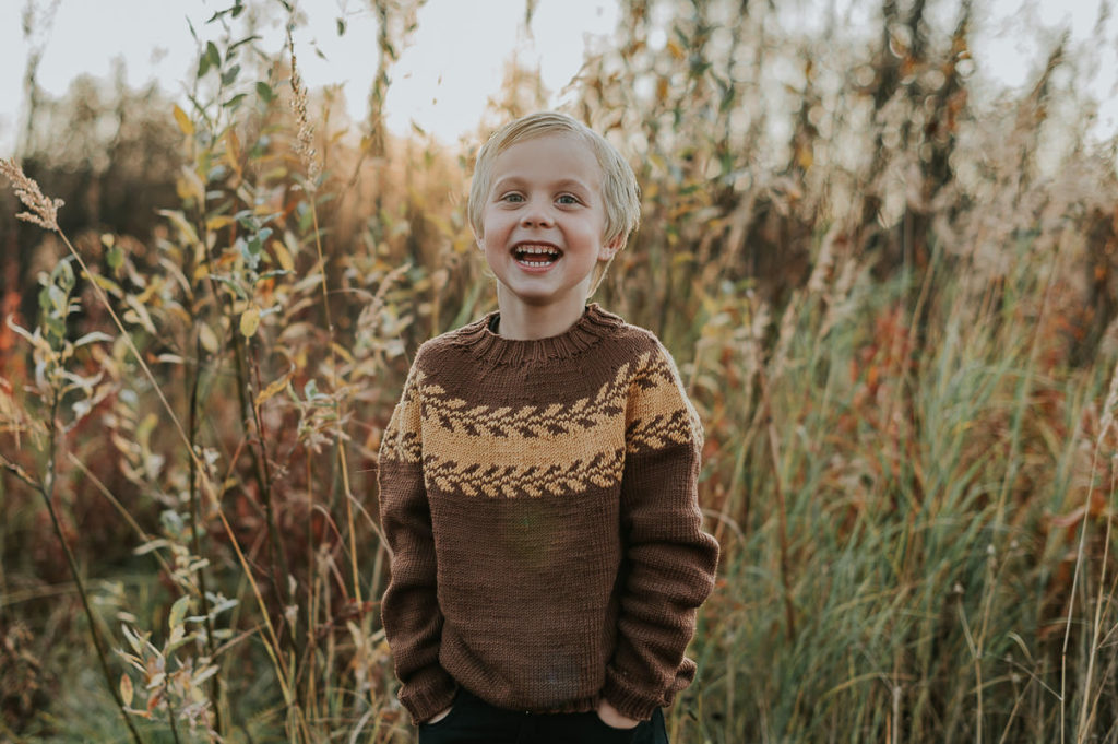 Barnefotografering i Alta blant fine høstfarger - en fin gutt i en strikket genser smiler til fotografen med nydelig  solnedgang i bakgrunn
