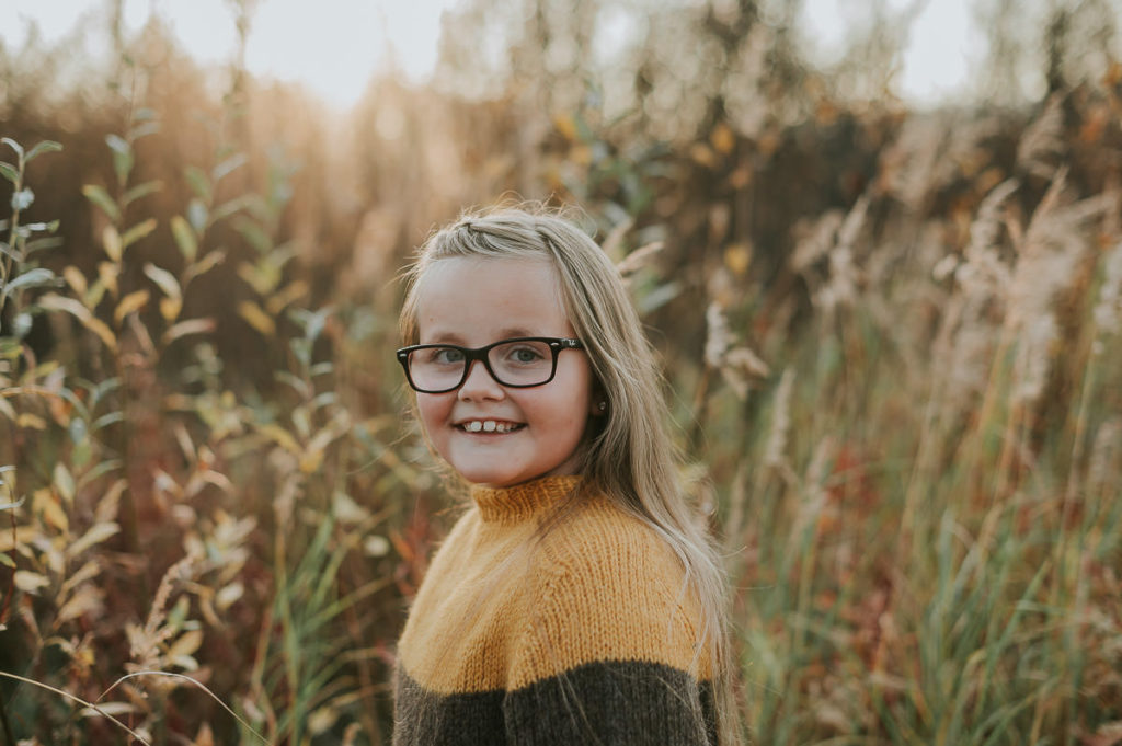 Barnefotografering i Alta blant fine høstfarger - en fin jente i en strikket genser smiler til fotografen med nydelig  solnedgang i bakgrunn