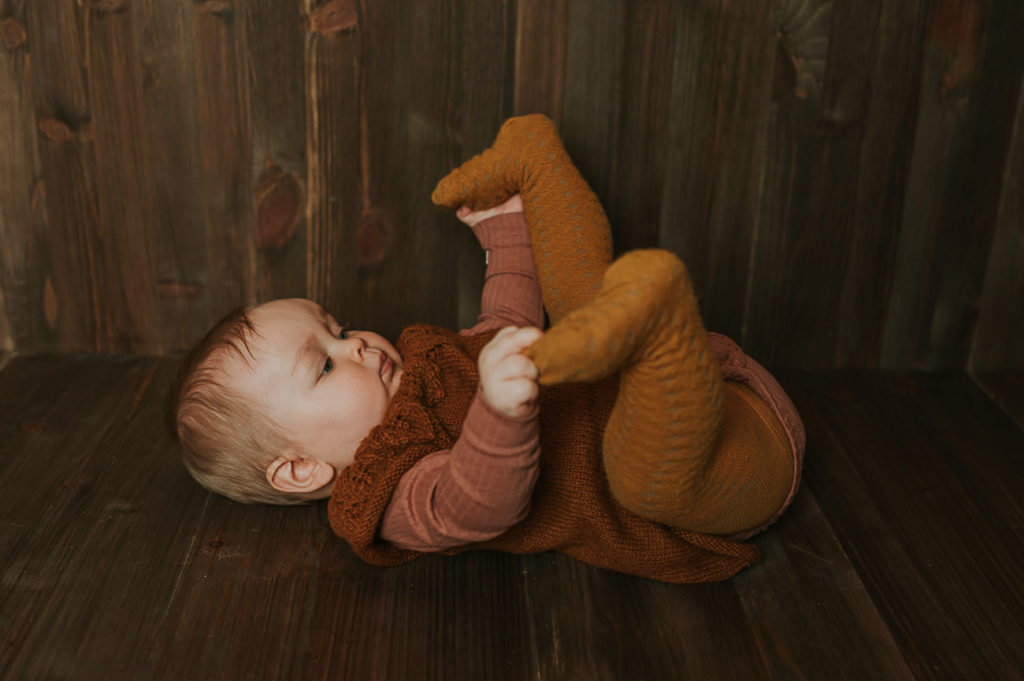 En 8 måneder gammel baby ligger på brunt rustikk fotobakgrunn og studerer føttene sine
