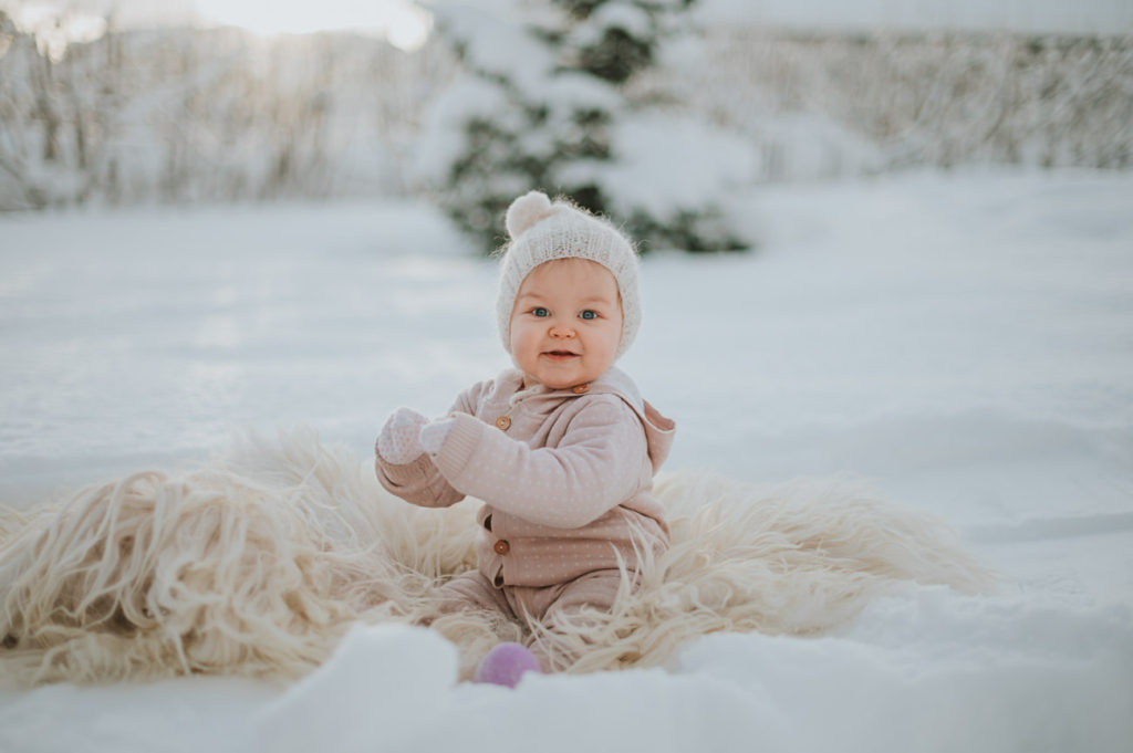 Nydelig jente på babyfotografering utendørs i Alta i flotte vinter omgivelser. Babyen sitter på sauskinn med soloppgang i bakgrunn og flott vinter landskap