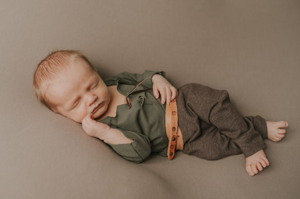 Cute newborn baby boy in a viking style outfit with a viking belt sleeping on a beige photo backdrop for his newborn photoshoot with viking theme