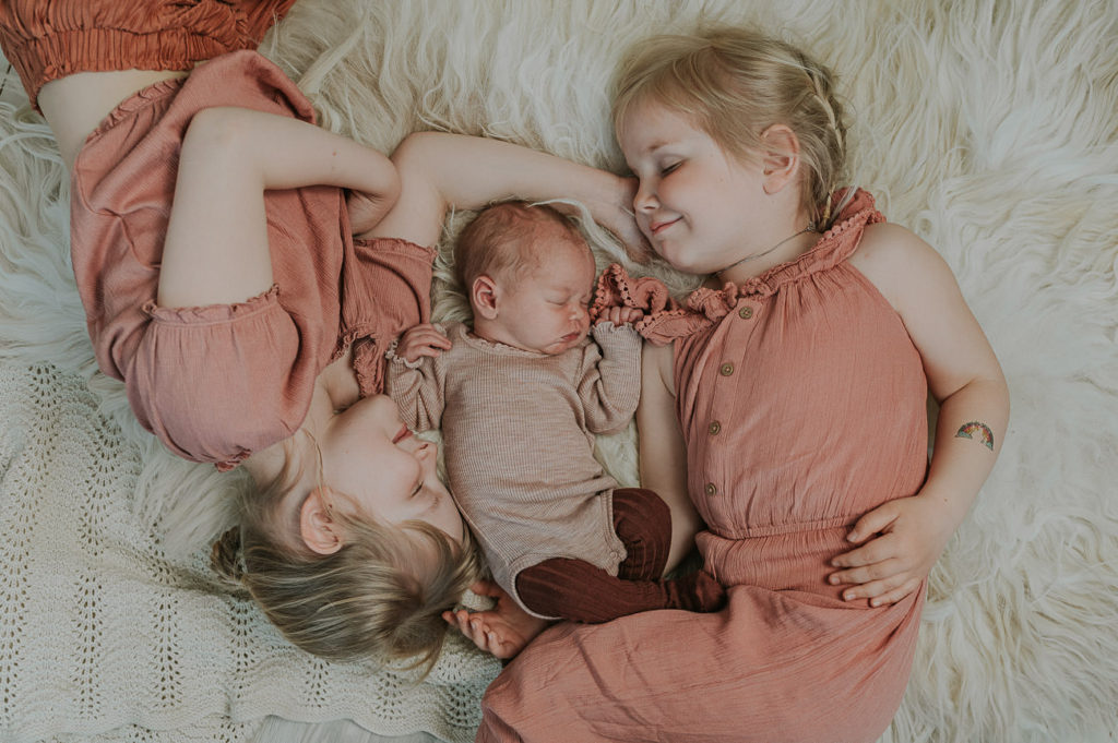 Søt nyfødt jente sammen med sine storesøster som ligger på sauskinn og smiler. Alle tre har rosa antrekk på seg