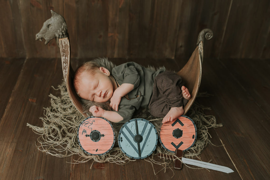 Viking theme newborn photoshoot in Norway. A baby in a viking outfit sleeping in a viking ship with Raagnar Lodbrok's shield and sword near him