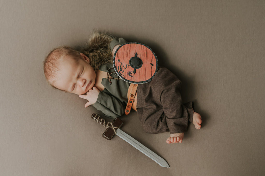 Cute newborn baby boy in a viking style outfit with a viking belt sleeping on a beige photo backdrop for his newborn photoshoot with viking theme. The baby is holding a shield and a sword inspired by tv show Vikings