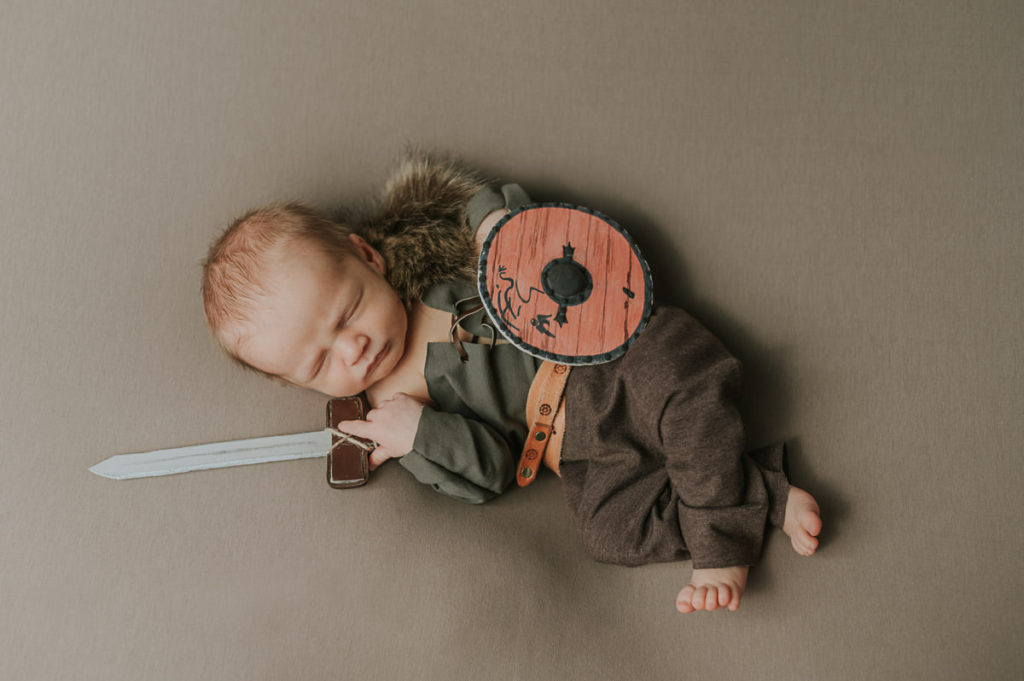 A cute newborn baby boy in a viking outfit with a sword and shield sleeping during viking theme photo session 