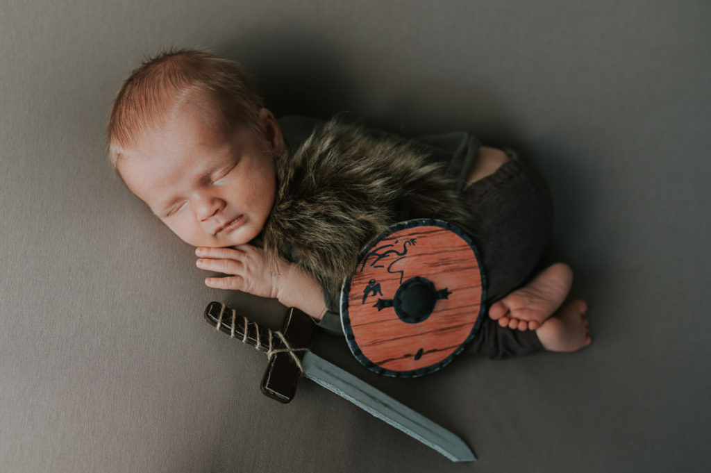 Sweet newborn boy in a  viking style outfit sleeping on a newborn photo session with a Ragnar Lodbrok sword and shield 