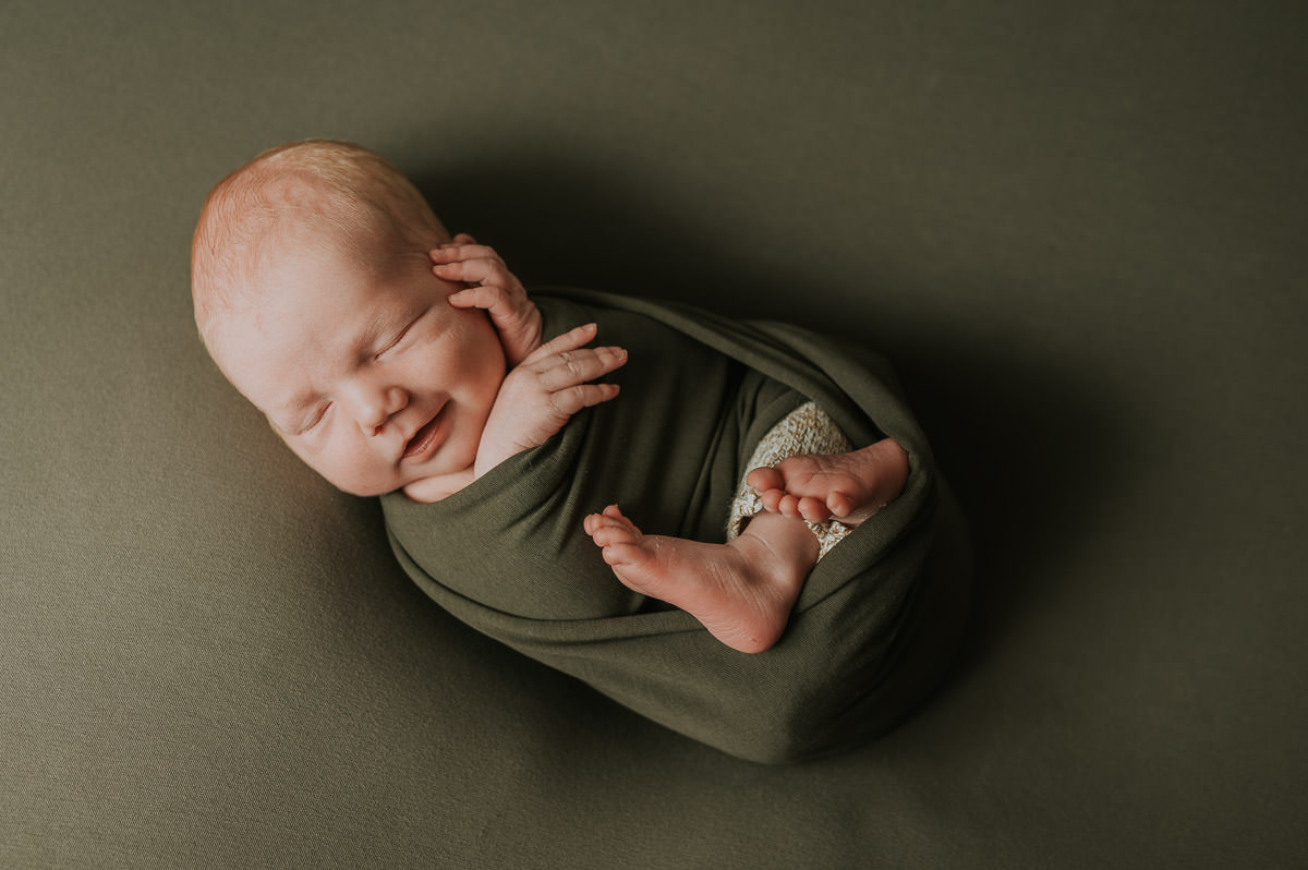 Nyfødt gutt tullet inn i grønn sjal på grønn fotobakgrunn smiler i søvne fotografert av TS Foto Design