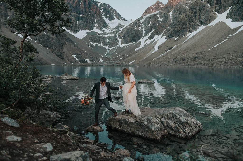 Groom helping the bride to walk on the rocks near a beautiful blue lake in Norway