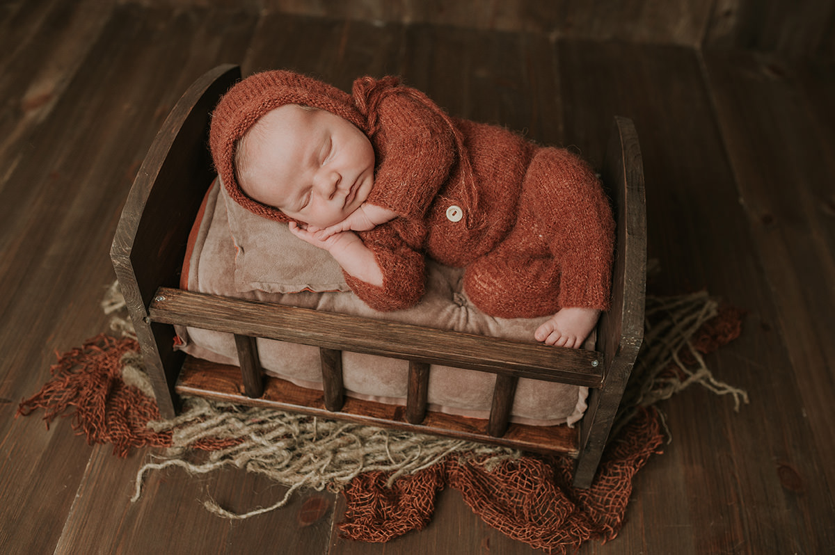 Nydelig gutt i fin rustfarget strikket dress og lue sover i en trebolle på nyfødtfotografering hos TS Foto Design