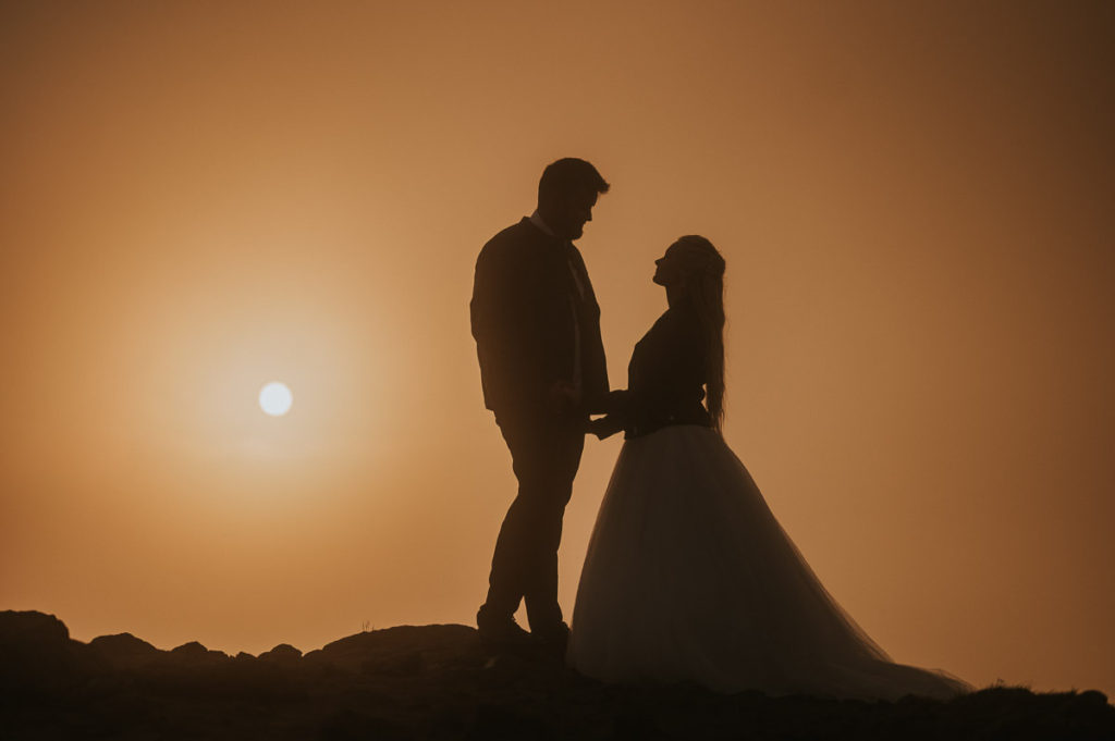 silhouette portrait of a bride and groom laughing and kissing each other in the sunset while clouds cover the sun on a mountaintop in Lofoten. The couple is enjoying their adventure elopement wedding day