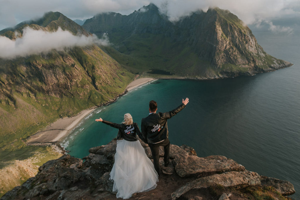 Mountain adventure elopement wedding in Lofoten islands - bride and groom wearing their wedding attire and leather jackets standing on a rock on a mountaintop and enjoying mesmerising views