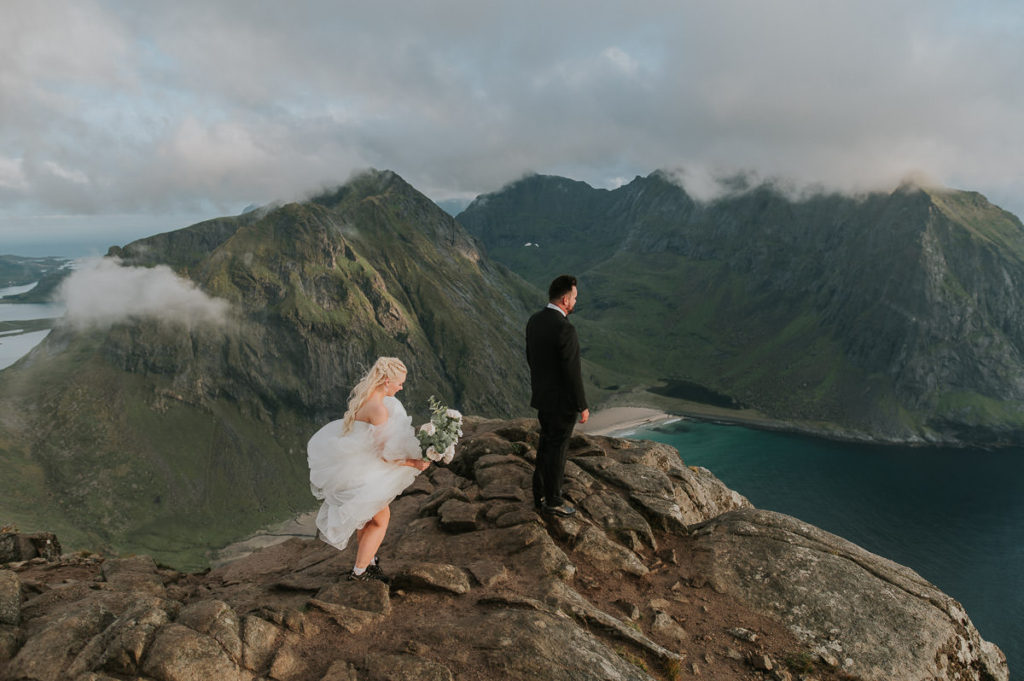 First look for bride and groom - mountain adventure wedding in Lofoten. The groom is waiting on a mountaintop for his bride while enjoying stunning view to a beach and mountains