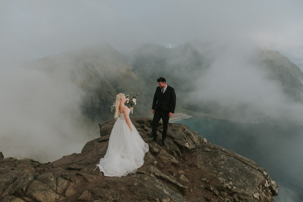 First look for bride and groom - mountain adventure wedding in Lofoten. The groom is waiting on a mountaintop for his bride while enjoying stunning view to a beach and mountains