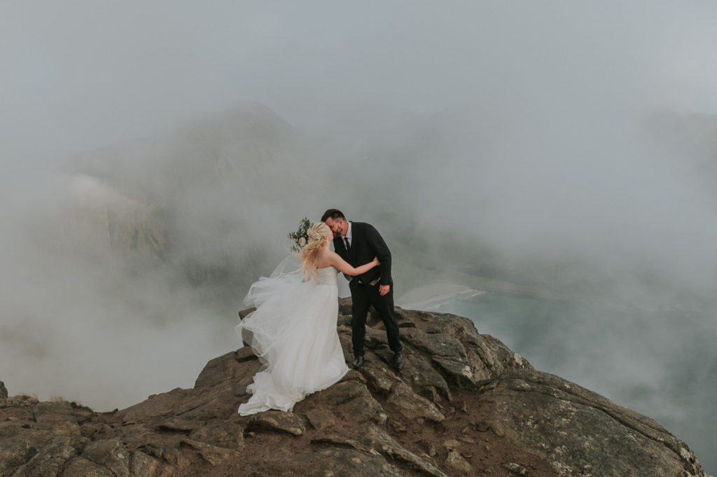 First look for bride and groom - mountaintop adventure wedding in Lofoten. Bride and groom kissing each other while clouds and fog rolls in