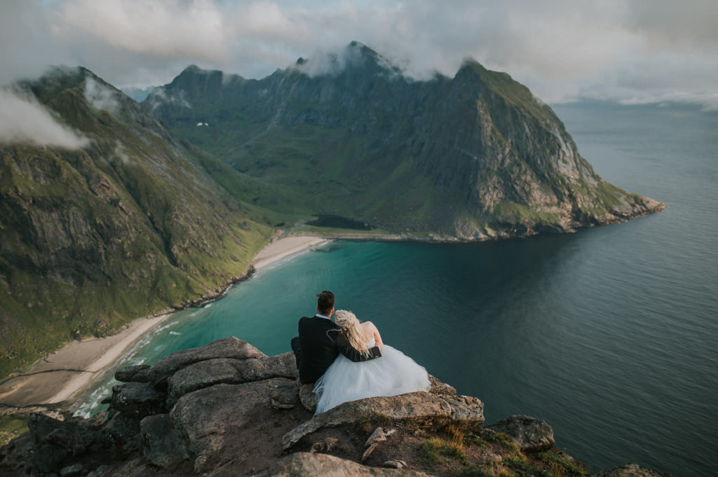 Bridal portraits on a mountaintop in Lofoten islands in Norway - bride and groom are sitting on a rock overlooking beautiful views to Kvalvika beach and stunning mountains