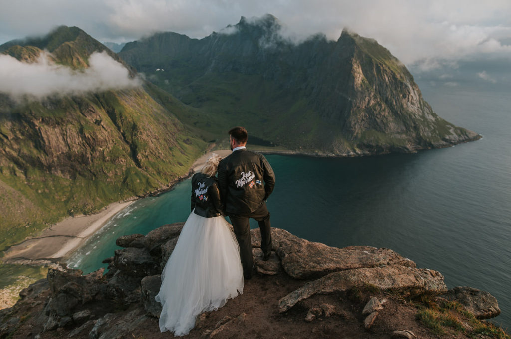 Mountain adventure elopement wedding in Lofoten islands - bride and groom wearing their wedding attire and leather jackets standing on a rock on a mountaintop and enjoying views