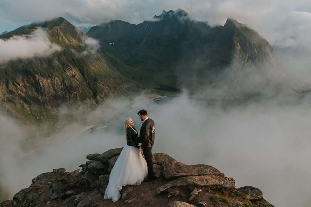 Mountain adventure elopement wedding in Lofoten islands - bride and groom wearing their wedding attire and leather jackets standing on a rock on a mountaintop and enjoying views
