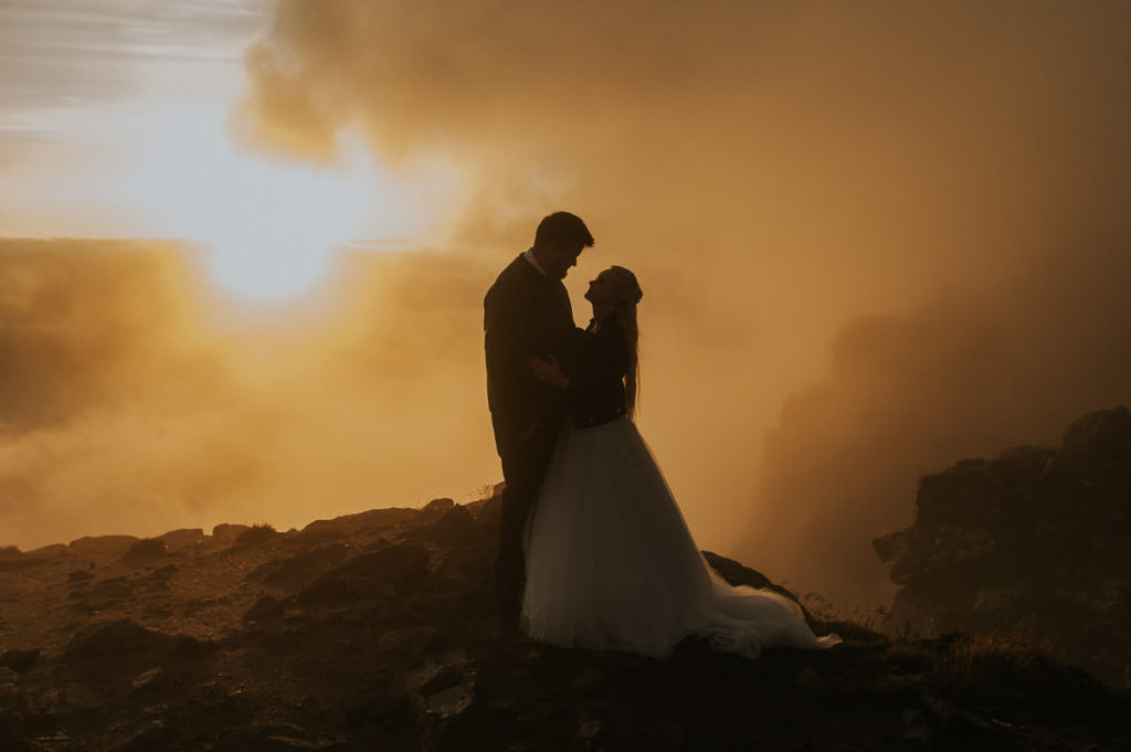silhouette portrait of a bride and groom kissing each other in the sunset while clouds cover the sun on a mountaintop in Lofoten. The couple is enjoying their adventure elopement wedding day