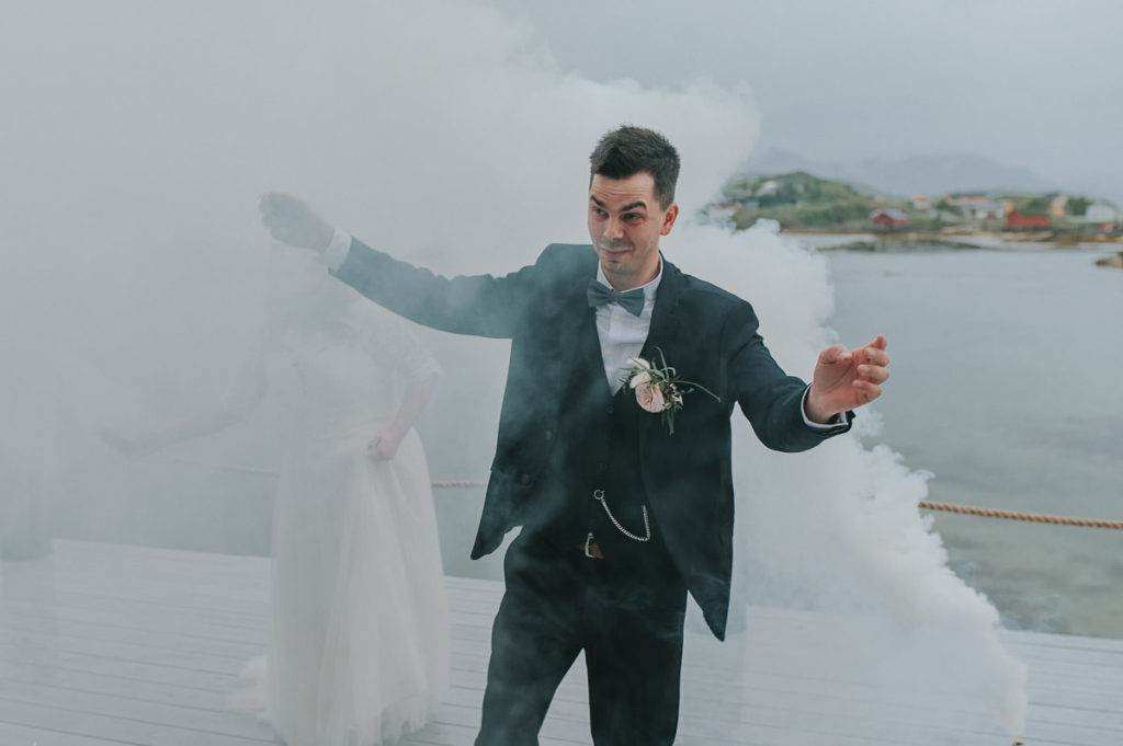 Bride and groom struggling to fire up smoke bombs while having funny expressions on their faces. The bride is hidden in the thick smoke 