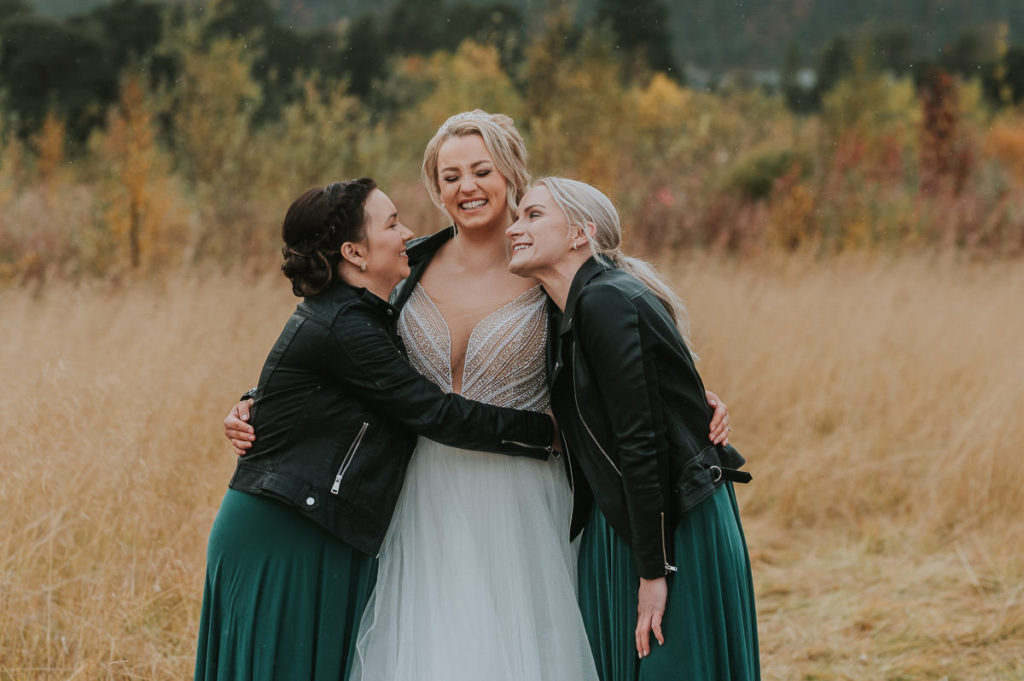 Bruden klemmer sine forlovere ikledd mørkegrønne kjoler og skinnjakker mens alle tre flirer. Bryllupsfotografering i Alta på en septemberbryllup