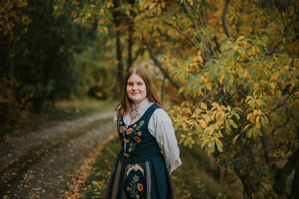 Portrett av ei jente i nordlandsbunad foran høstkledd skog på konfirmantfotografering i Alta