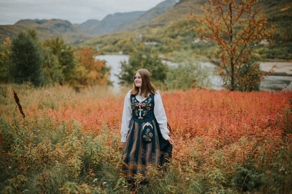 Portrett av ei jente i nordlandsbunad foran høstkledd skog og havet på konfirmantfotografering i Alta