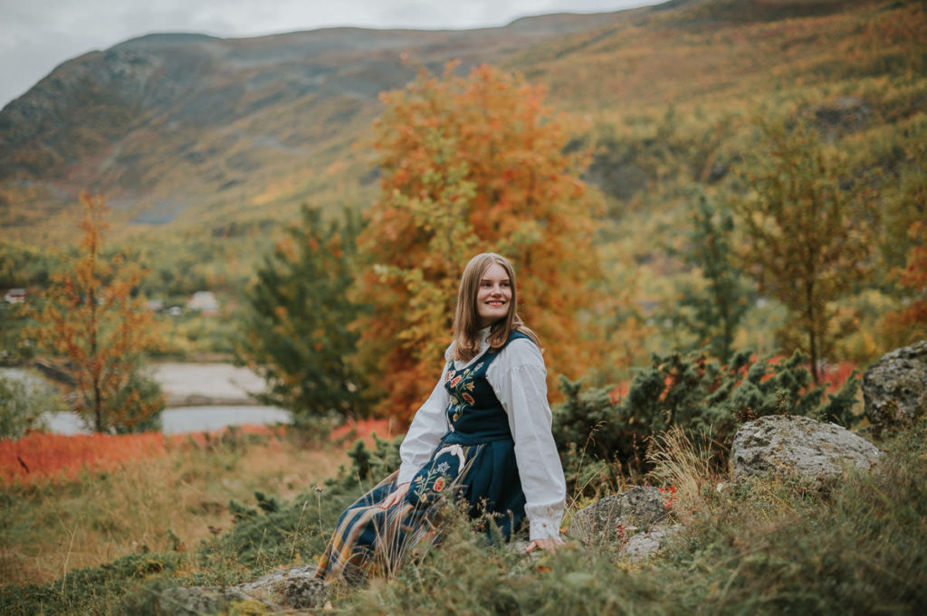 Portrett av ei jente i nordlandsbunad foran høstkledd skog og havet på konfirmantfotografering i Alta