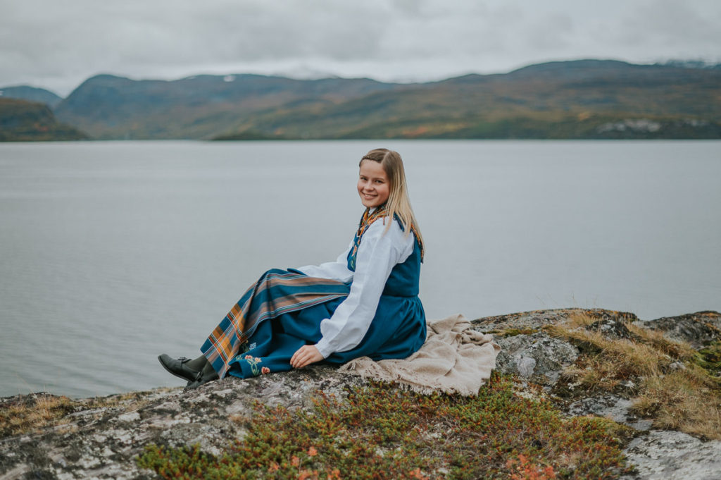 Portrett av ei jente i nordlandsbunad som sitter på bergene foran sjø i Tollevika i Alta på konfirmantfotografering