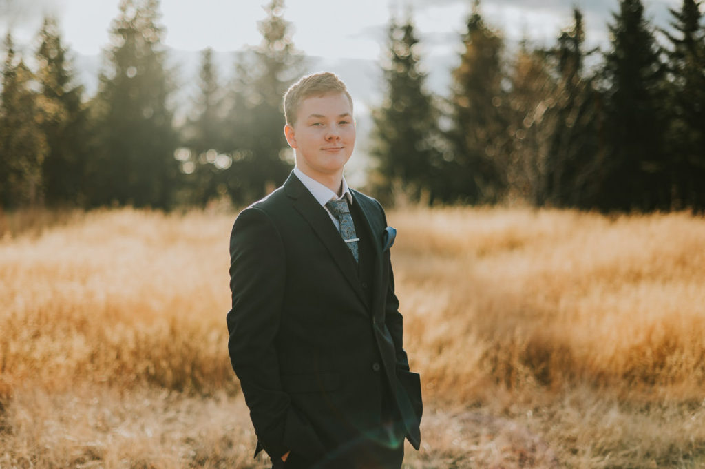 En konfirmant gutt kledd i dress poserer for konfirmantfoto blant nydelige høst omgivelser - et stråfelt og skog i Alta