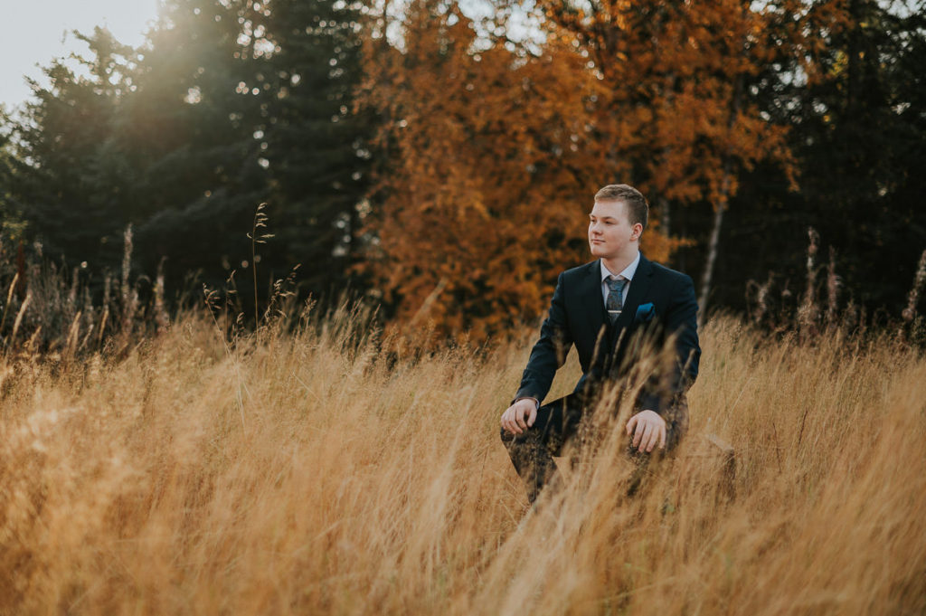 En konfirmant gutt kledd i dress poserer for konfirmantfoto blant nydelige høst omgivelser - et stråfelt og skog i Alta
